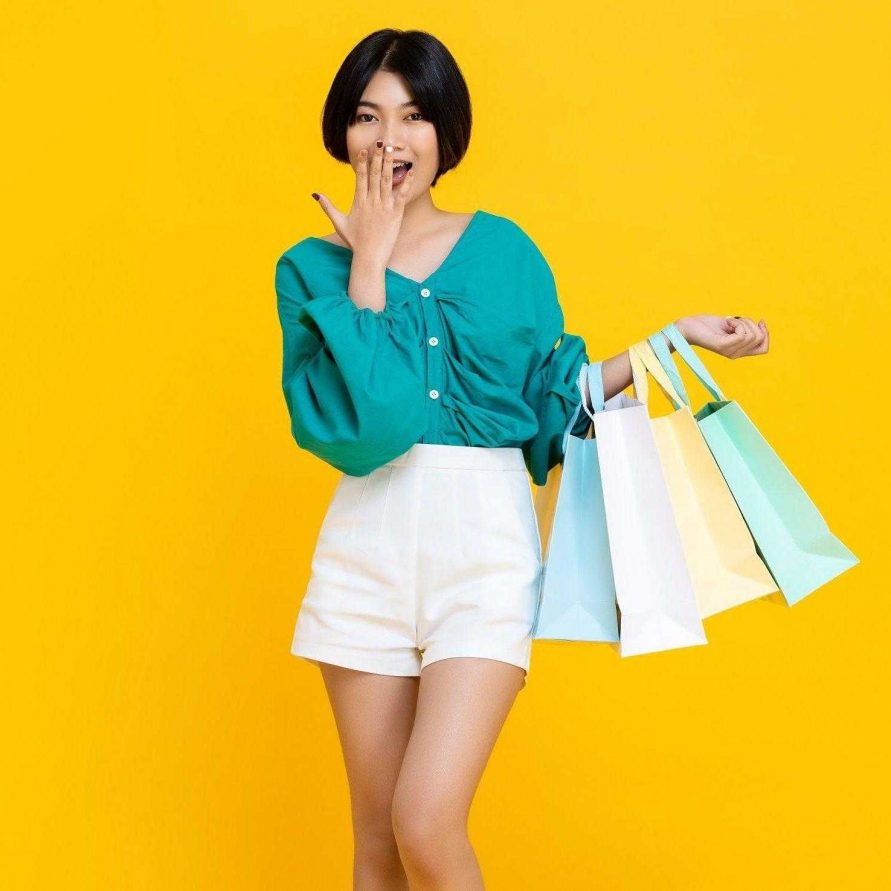 Young cheerful shopaholic Asian girl with lots of shopping bags on her arm isolated on yellow studio background with copy  space